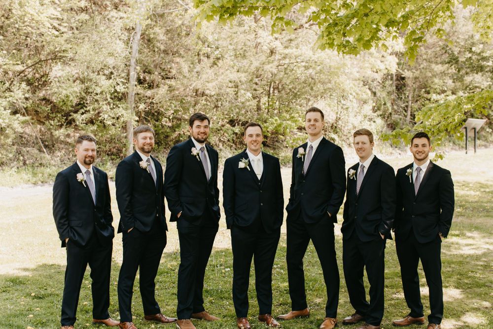 Seven groomsmen in navy suits with boutonnieres posing casually outdoors at wedding venue.
