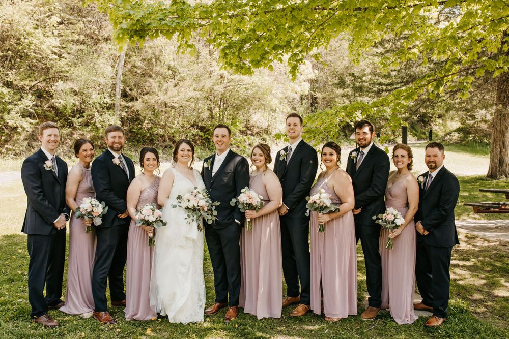 Wedding party poses outdoors in lush green park, bridesmaids in blush pink gowns and groomsmen in dark suits flanking bride and groom.