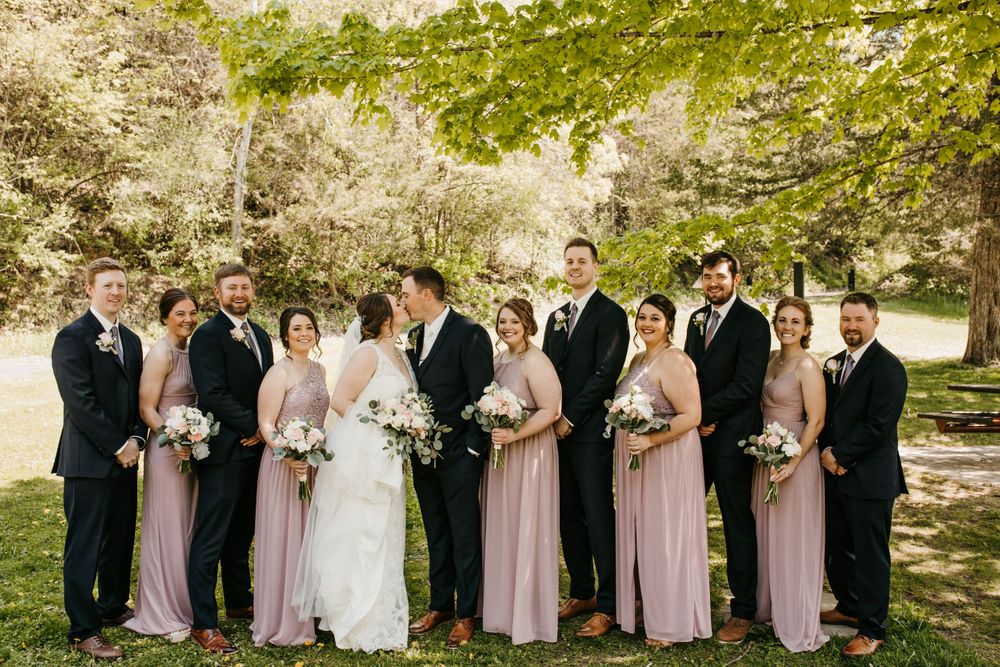 Wedding party shares a joyful moment kissing and laughing outdoors, bridesmaids in dusty rose dresses with white bouquets.