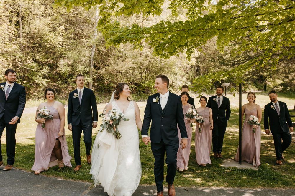 Bride and groom walk hand in hand with wedding party following through a sunny park, bride carrying large white floral bouquet.