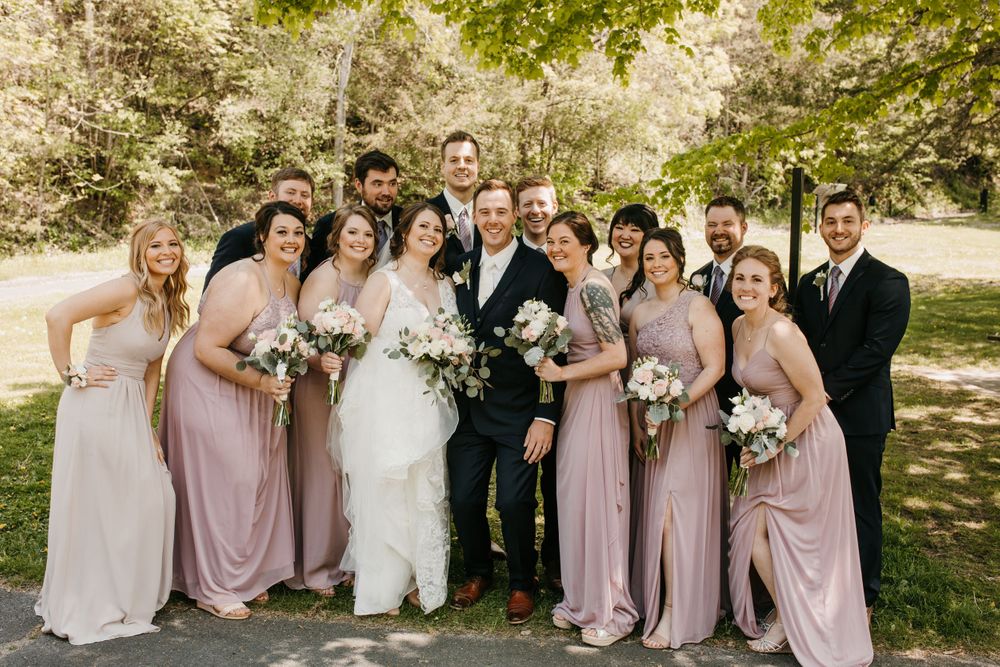 Wedding party gathers for a candid laughing group photo outdoors, bridesmaids in blush pink dresses holding white bouquets.