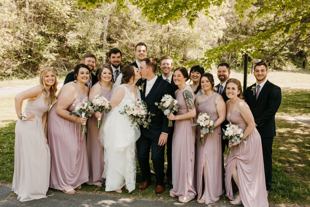 Smiling wedding party poses together under leafy green trees, bridesmaids in dusty rose gowns flanking bride in white dress.