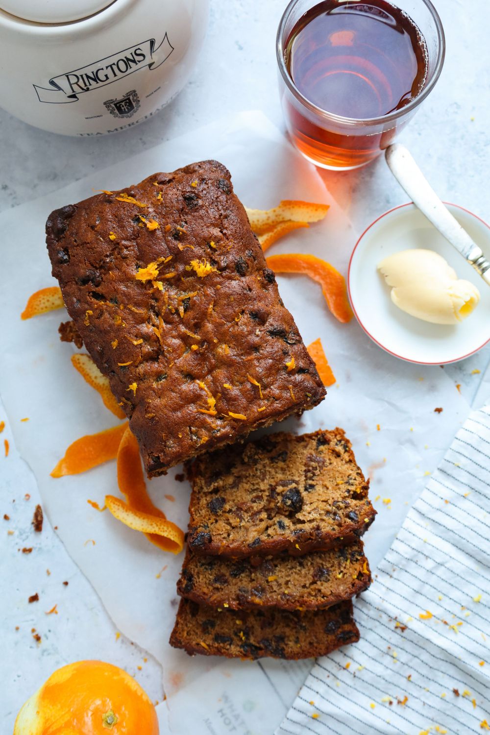 Slices of moist fruit cake with orange zest and tea served on a white surface.