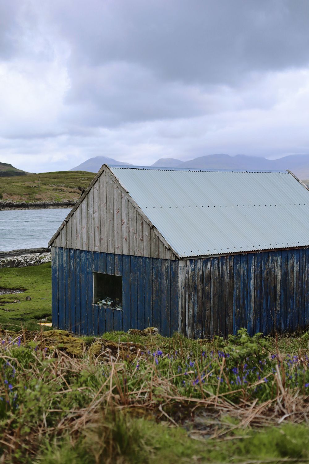 A weathered blue wooden barn with a metal roof stands in a rugged coastal landscape under cloudy skies.
