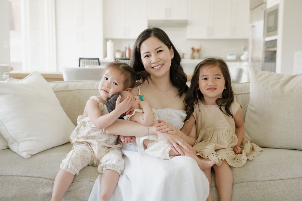 A family lounges together on a white couch in coordinated neutral-toned outfits for a casual home photoshoot.
