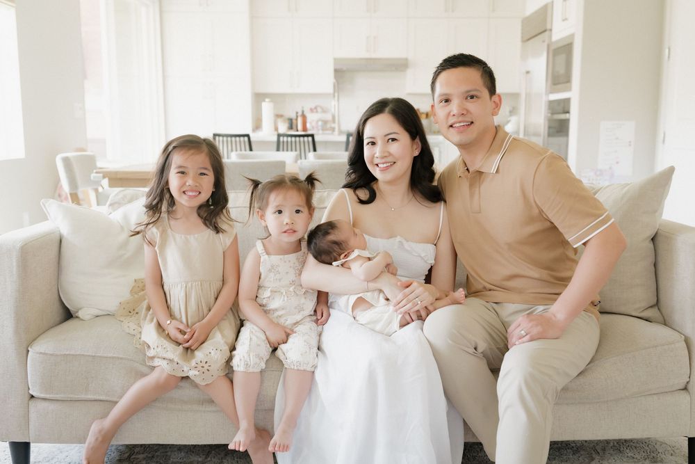 A family sits together on a light beige couch in a bright, airy living room wearing coordinated neutral outfits.