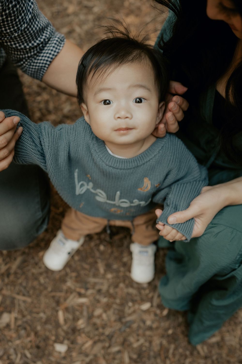 A small child wearing a blue sweater and dark pants sits on fallen leaves in an autumn outdoor setting.