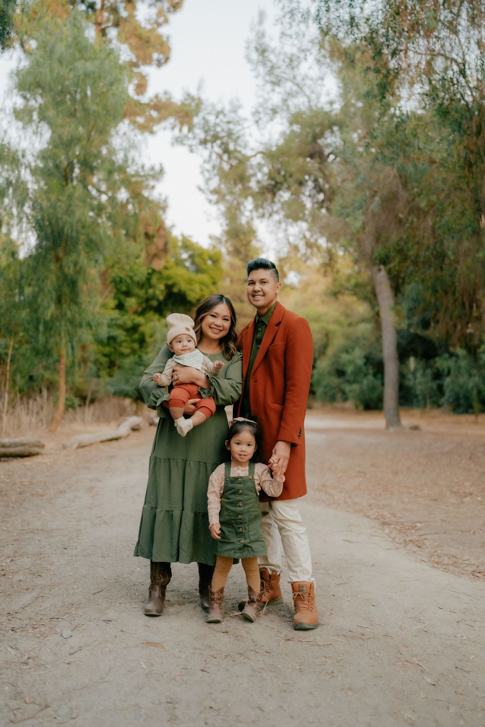 A family group stands together on a dirt path surrounded by trees during an autumn outdoor photo session.