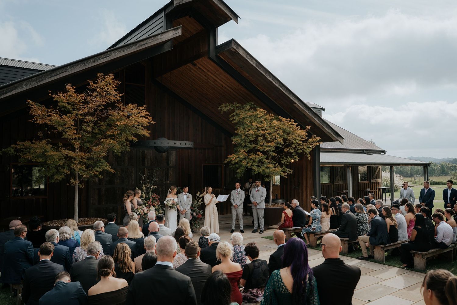Outdoor wedding ceremony with guests seated before a modern dark wood venue surrounded by autumn trees.