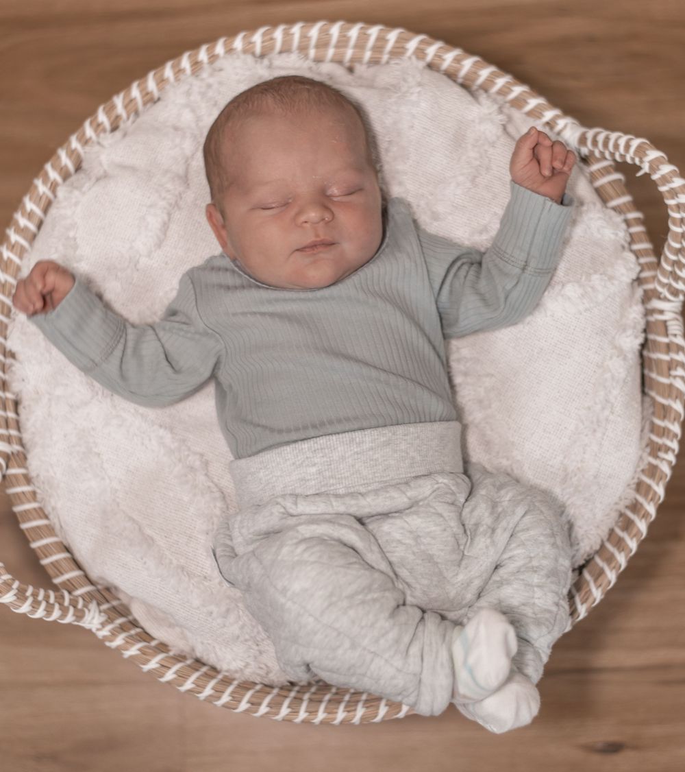 A sleeping newborn baby wearing a soft gray outfit lies in a cozy white basket on a wooden floor.