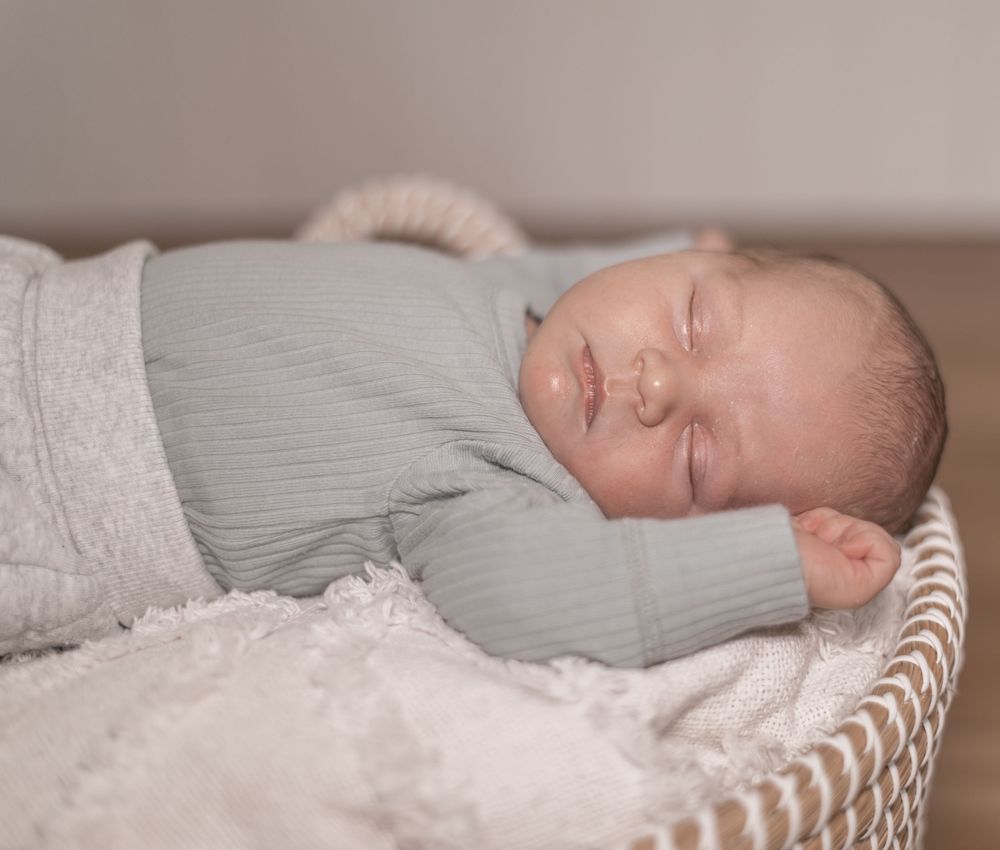 A peaceful sleeping newborn baby wrapped in soft gray fabric rests in a white woven basket.