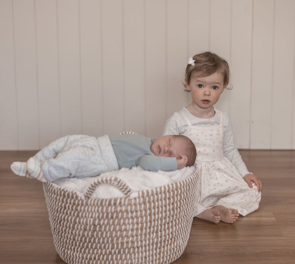 A baby rests peacefully in a woven basket while wearing a white outfit.