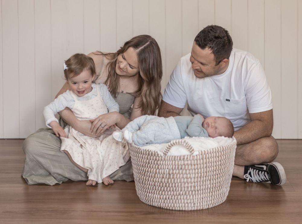 A family sits together on a hardwood floor with a baby in a woven basket while sharing a tender moment.