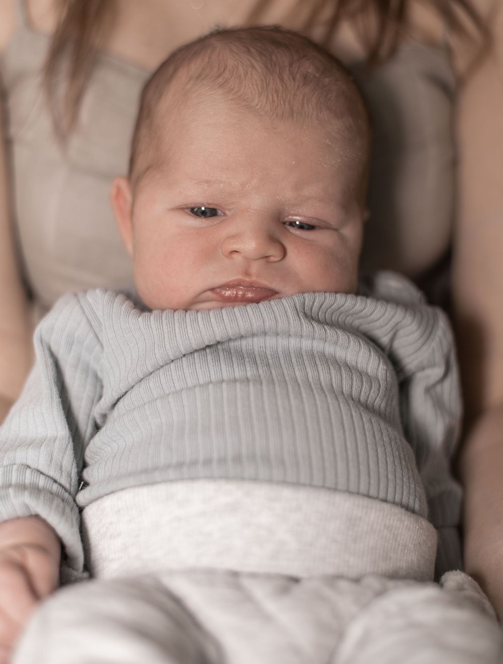 A newborn baby wearing a soft gray ribbed sweater lies peacefully on a light-colored blanket.
