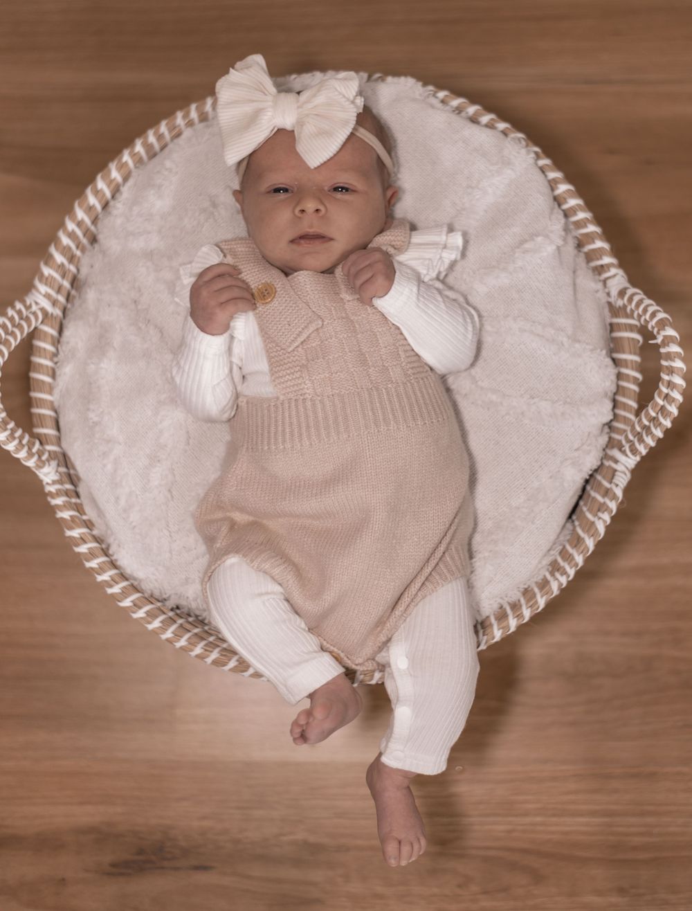Newborn baby wearing beige outfit lying on soft white blanket in rustic setting.