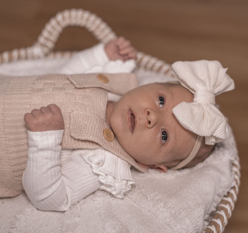 A baby in white christening gown lies on a cream-colored woven basket against a brown backdrop.