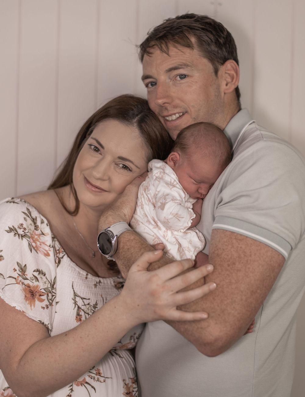 A couple holds their sleeping newborn baby while posing together against a beige wall.