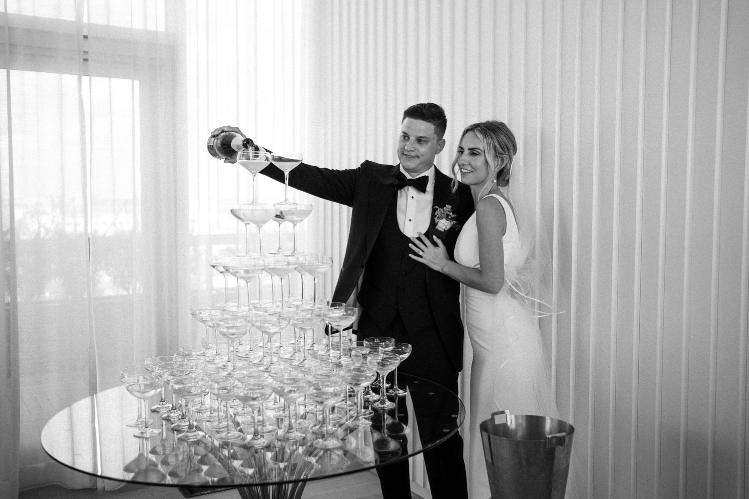 Wedding couple pouring champagne into tower of glasses at reception.