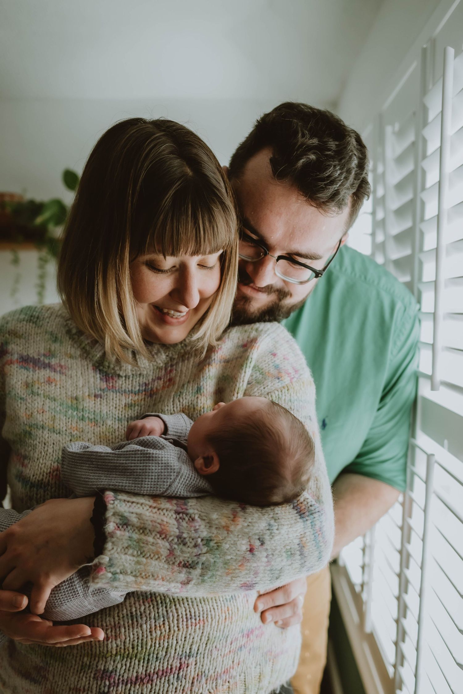 Parents share a tender moment while holding their newborn baby near a window with plantation shutters.