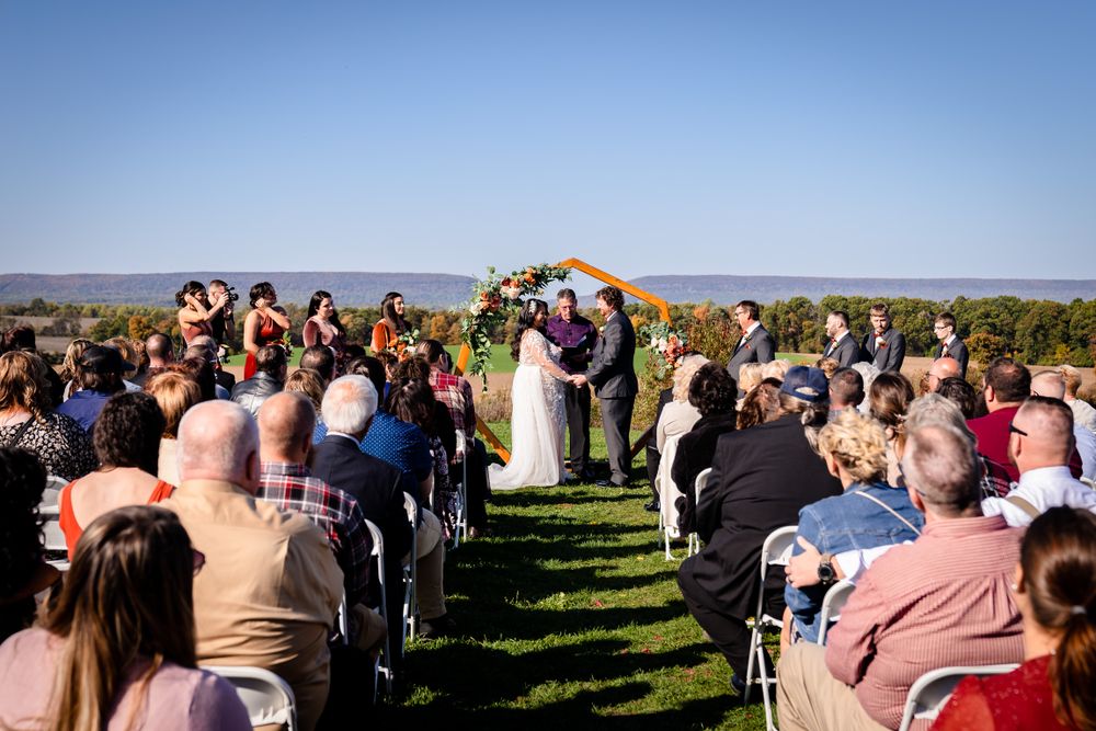 Outdoor wedding ceremony with guests seated in rows on a hillside overlooking scenic landscape.