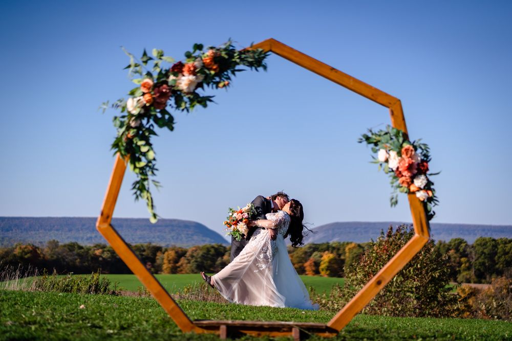 Wooden hexagonal wedding arch decorated with pink and white flowers against a bright blue sky.
