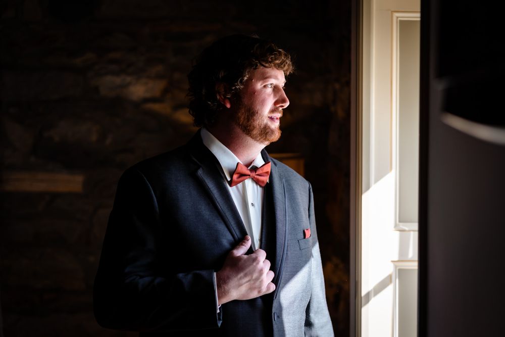 Groom adjusts his tie while getting ready in natural window light.