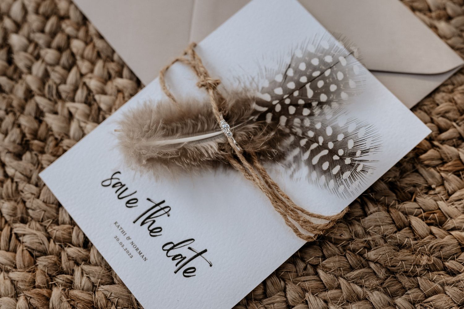 Wedding invitation card decorated with feathers and twine on woven surface.