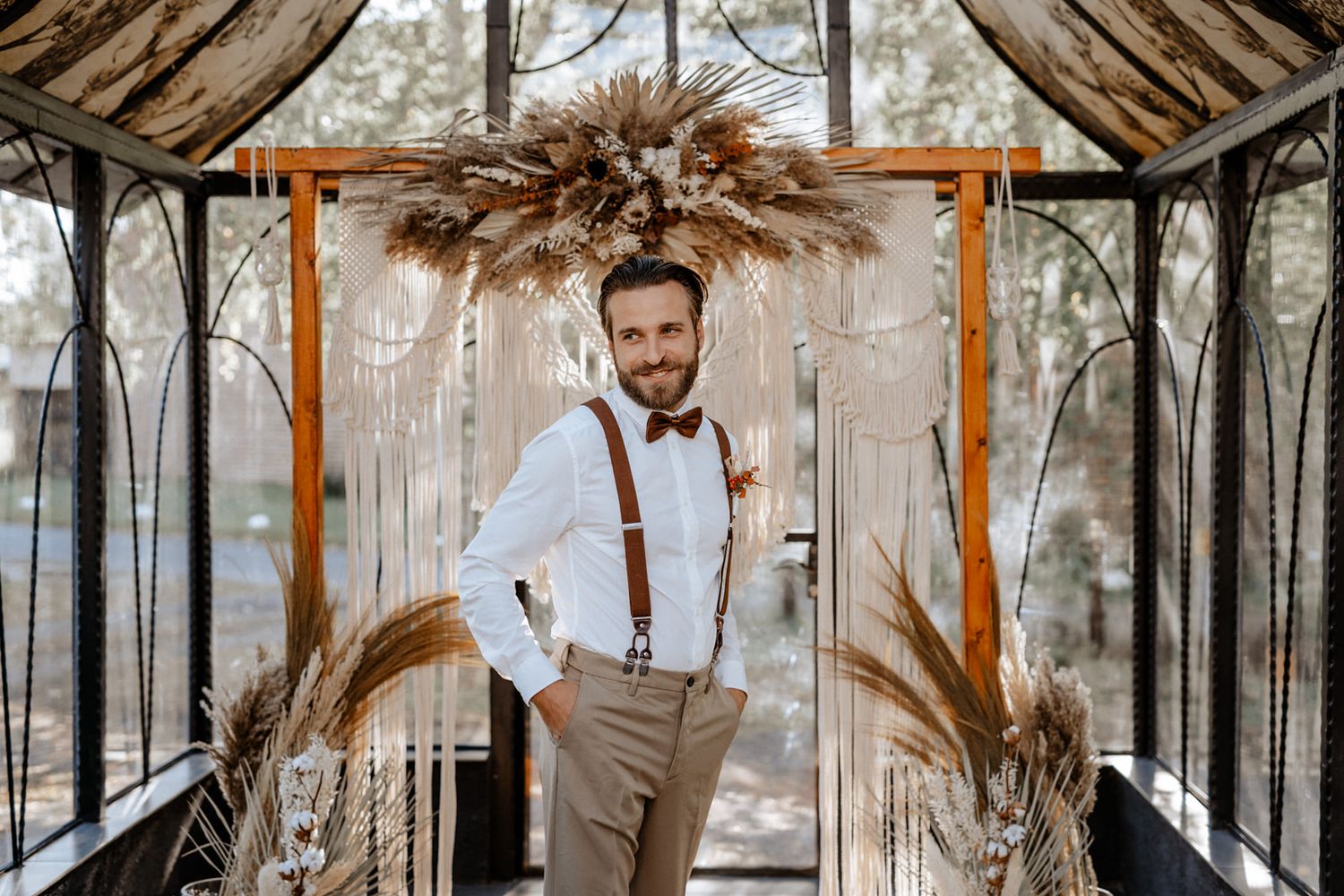 Groom in suspenders and bow tie standing under rustic wedding arch.
