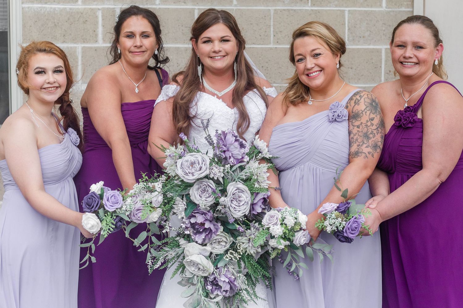 Wedding party members holding elegant gray and purple bouquets while posing together.