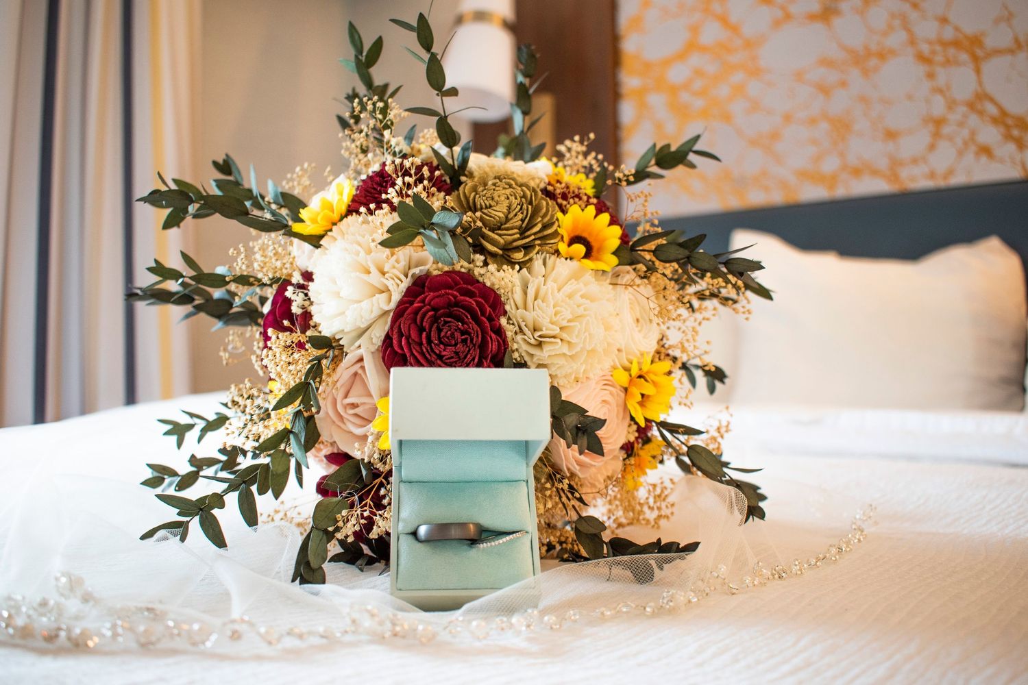Wedding bouquet with red and white flowers sits beside a turquoise ring box on white bedding.