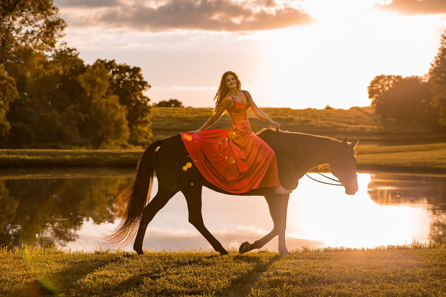 Beautiful girl in a red, flowing dress on a black horse, captured walking along a pond at sunset.