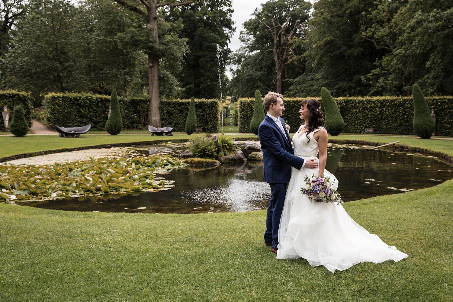 Wedding couple poses near ornate garden pond surrounded by manicured landscaping.
