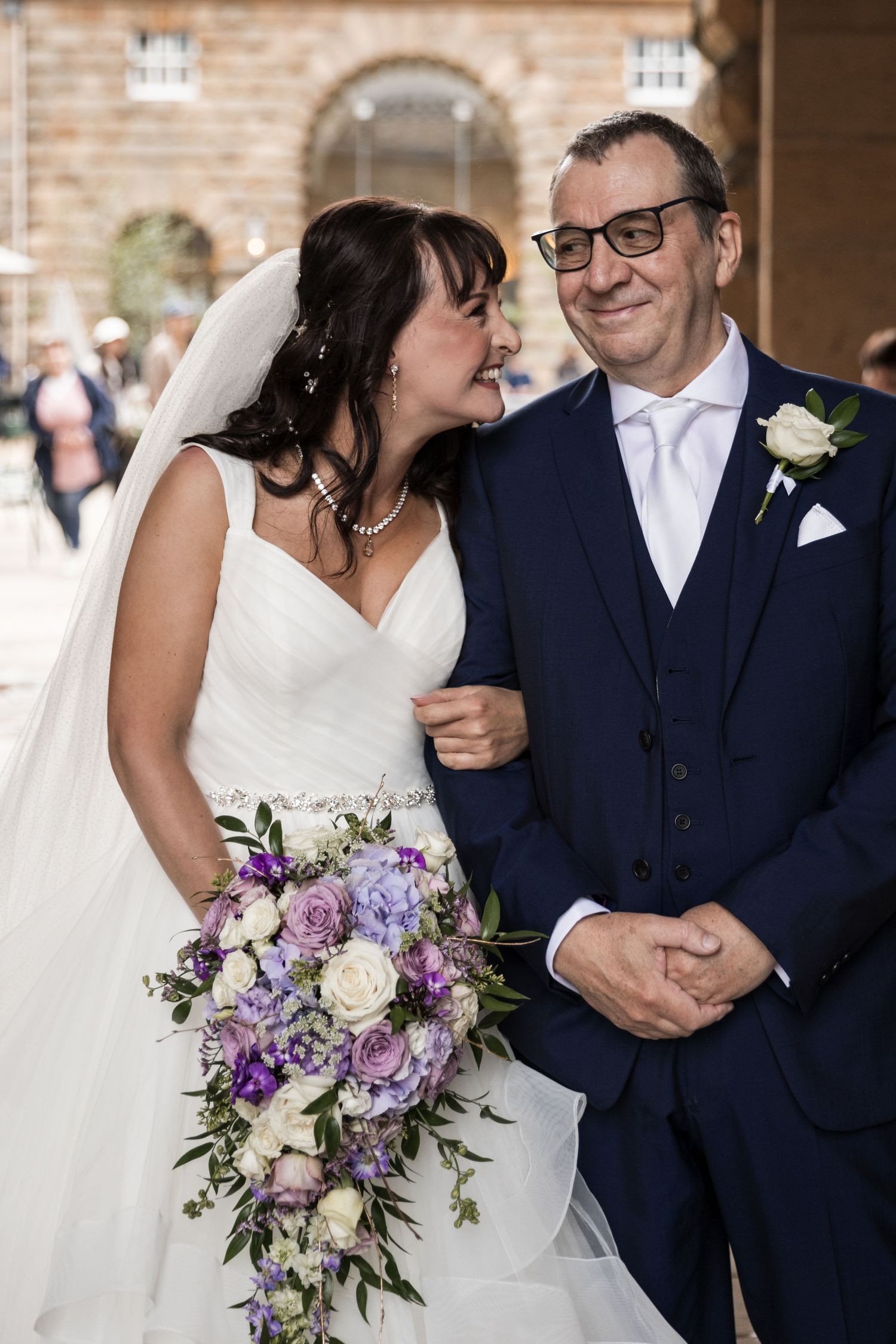 Wedding couple shares a joyful moment under brick archway with elegant floral bouquet.
