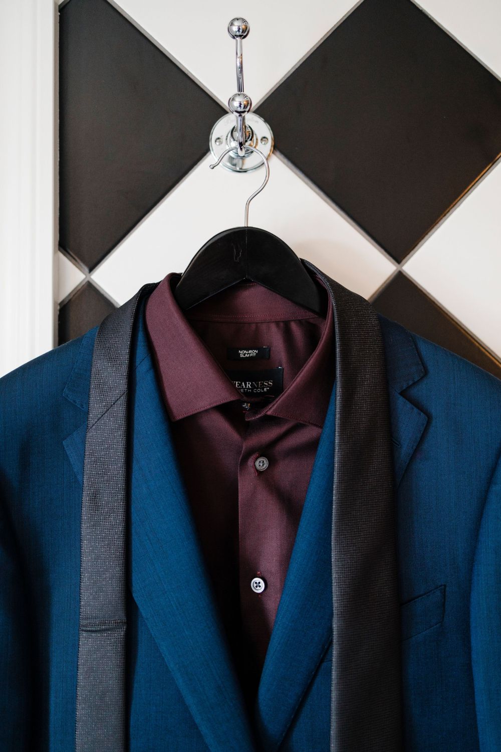 A blue suit jacket with burgundy shirt and gray suspenders hanging on black and white checkered wall.
