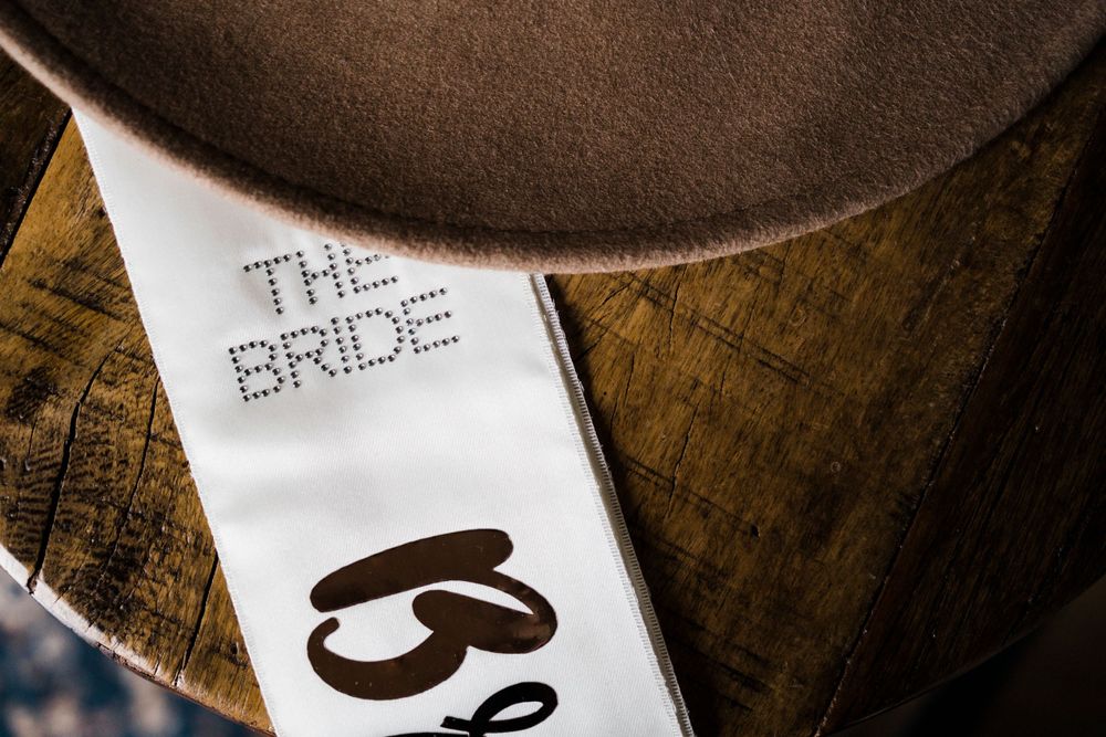 Close-up shot of a white sash with 'THE BRIDE' text in rhinestones against a wooden surface.