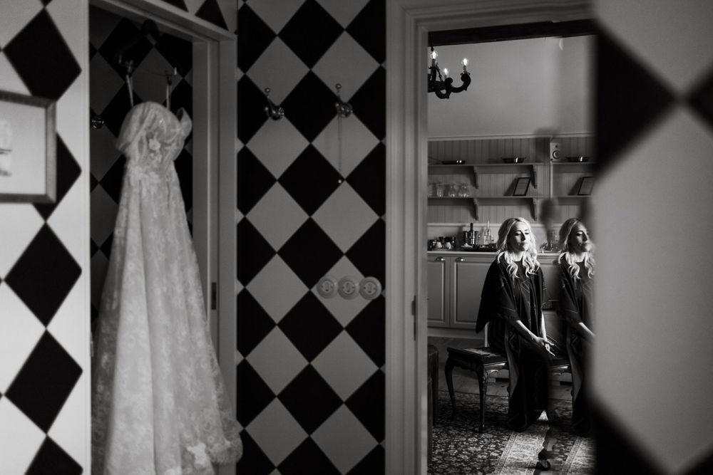 Black and white photo of a wedding dress hanging on checkered wall with room reflection in mirror.