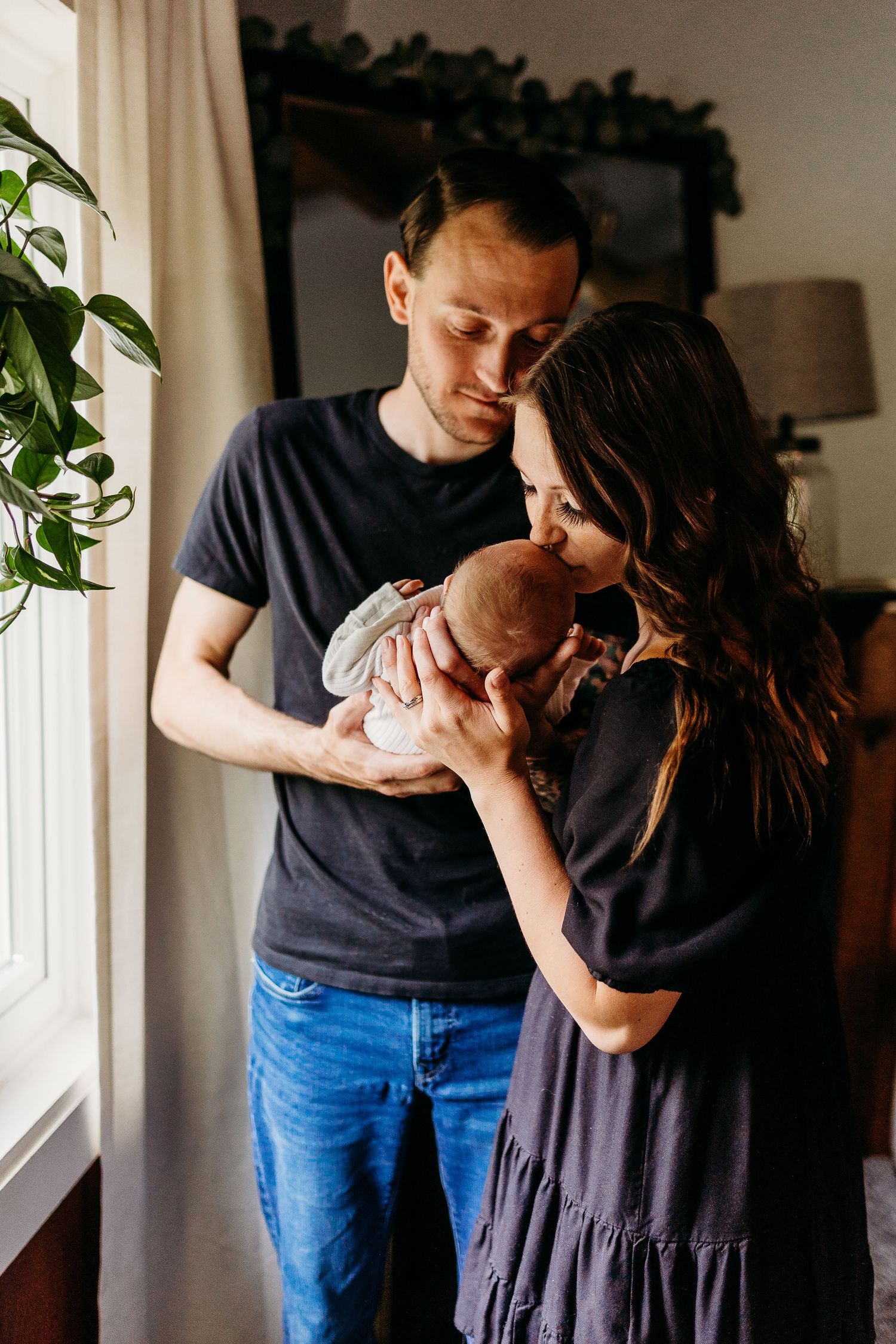 A couple shares an intimate moment with their newborn baby near a window with houseplants.