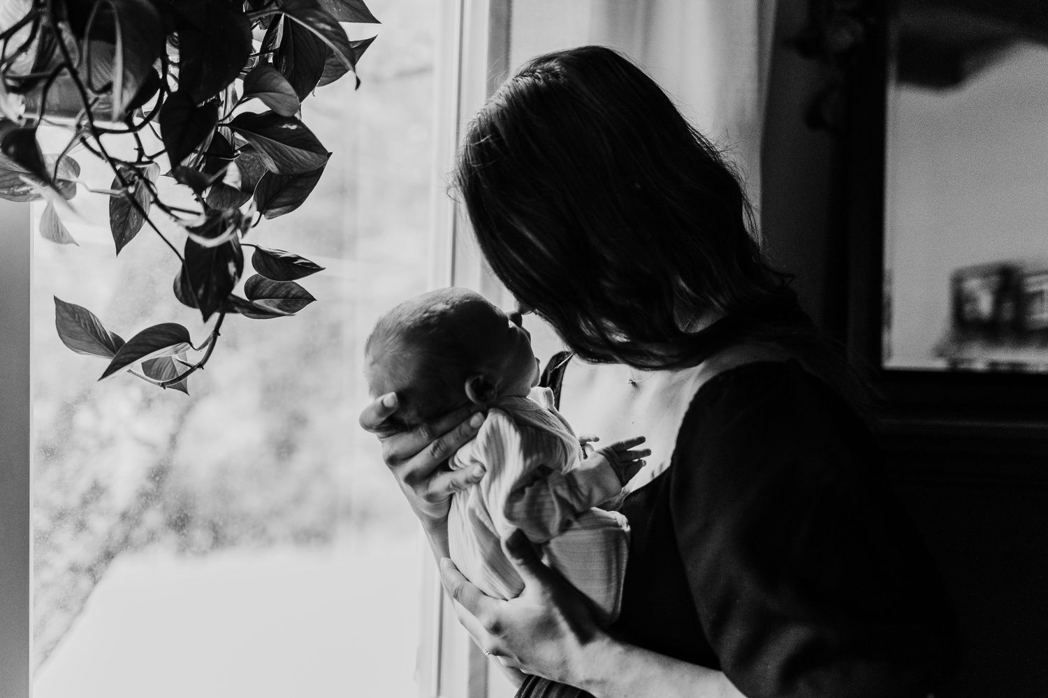 A black and white image of a parent cradling a small infant near a window with plants.