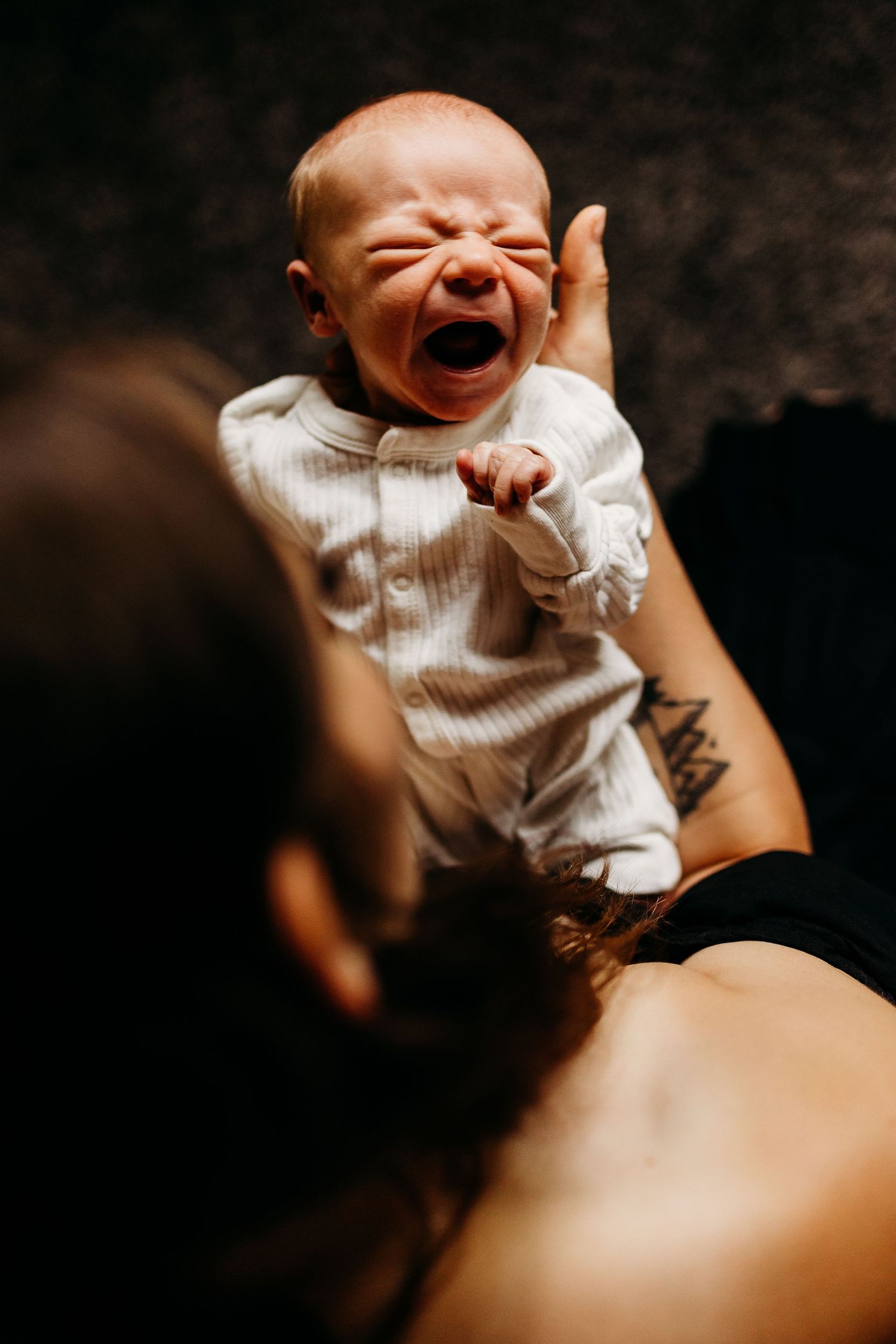 A crying newborn in white clothing is held up against a dark background.