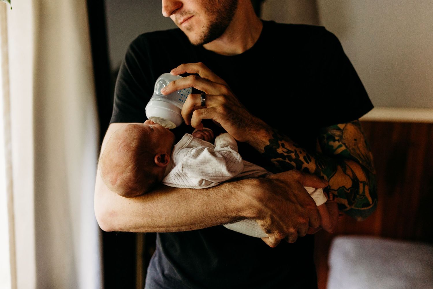 A person with sleeve tattoos cradles a newborn baby while standing in natural light.