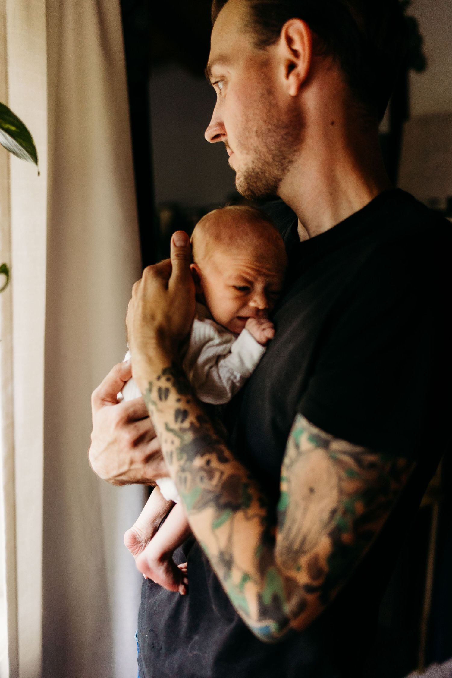 A person with tattooed arms holds a small infant while standing near a window with plants.