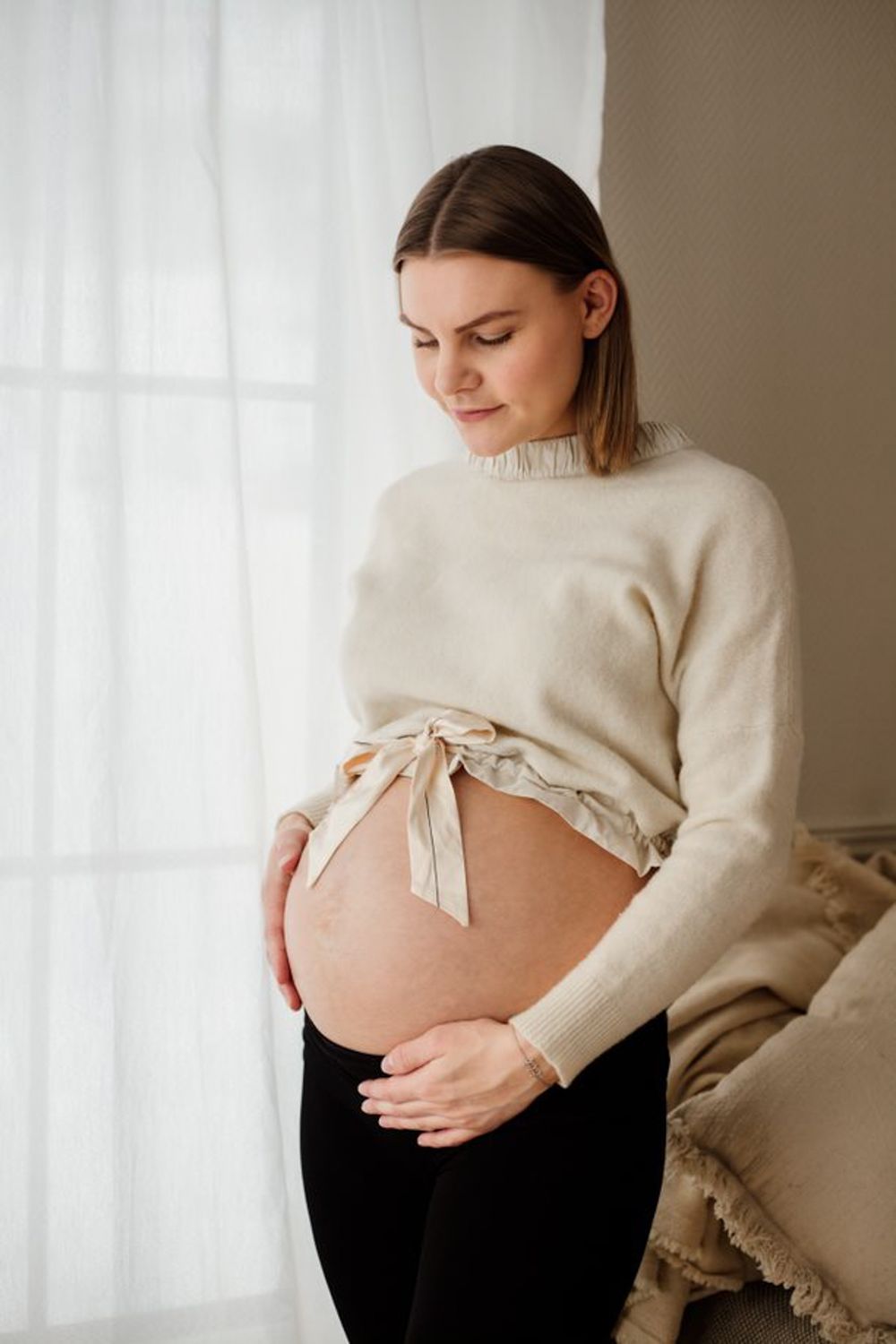 A maternity photo featuring a cream sweater and black pants against white curtains.