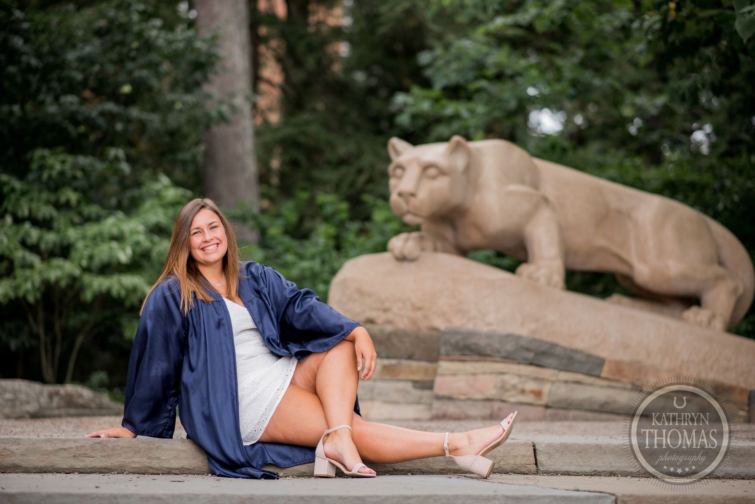 A graduate posing in casual attire next to a large stone lion sculpture on campus.