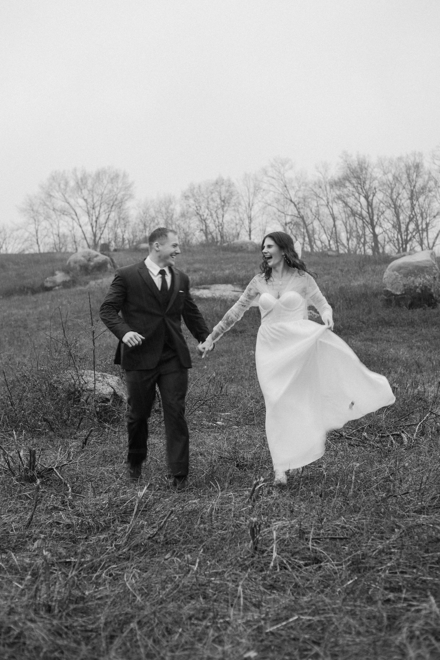 Couple in wedding attire running together across a rustic field.
