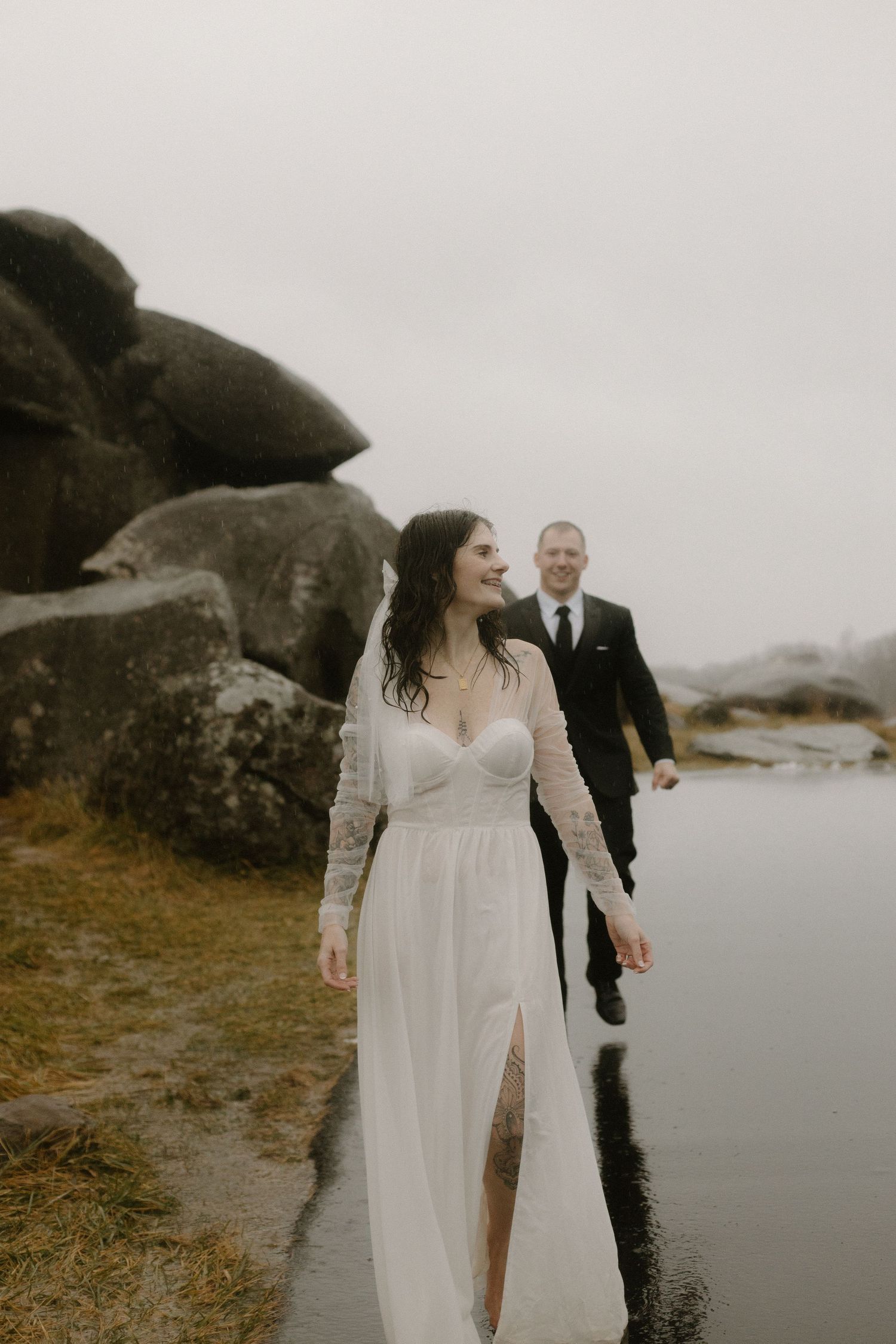 A couple in wedding attire walking near large boulders on a cloudy day.