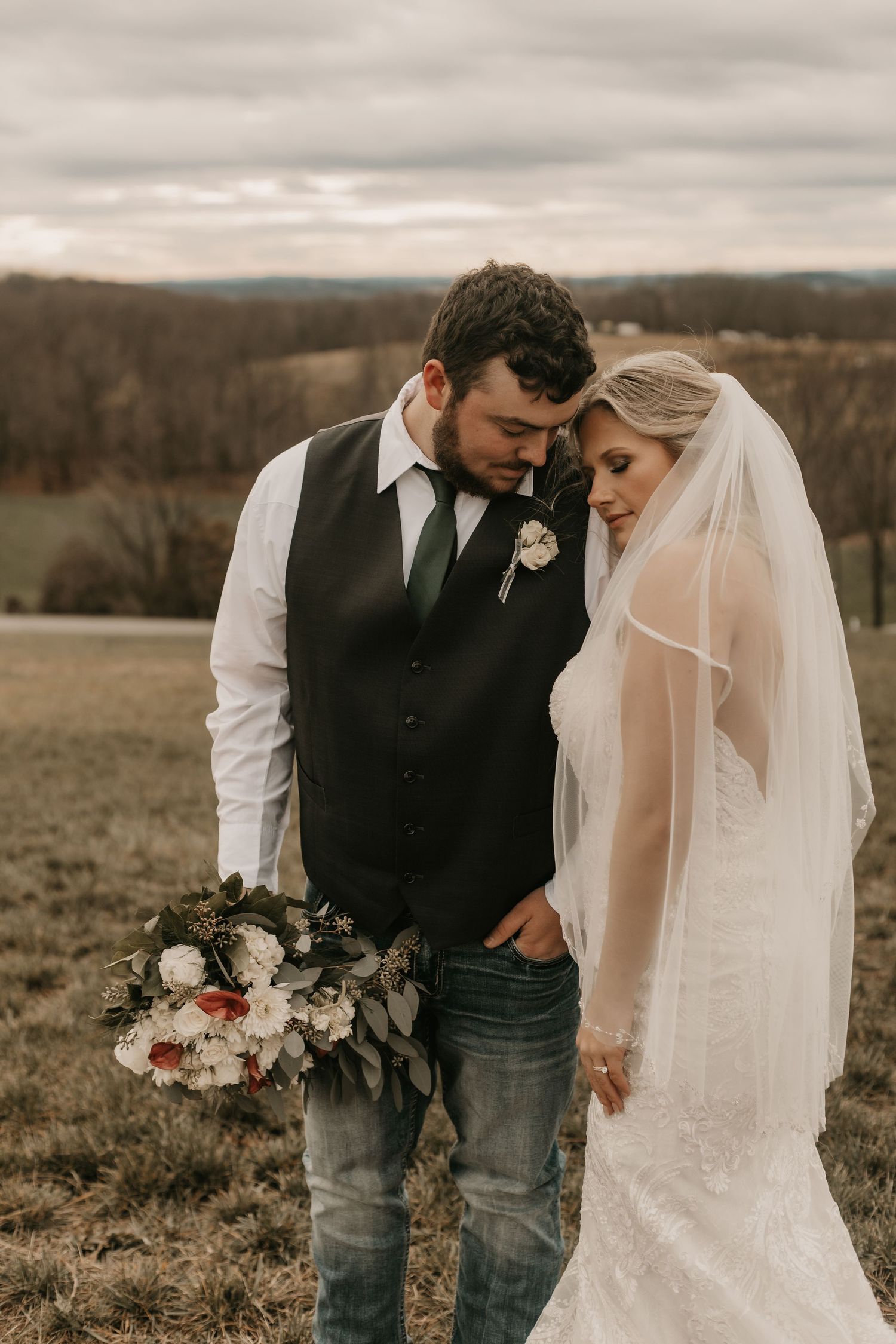 Romantic wedding portrait in a field with overcast sky and rolling hills in background.