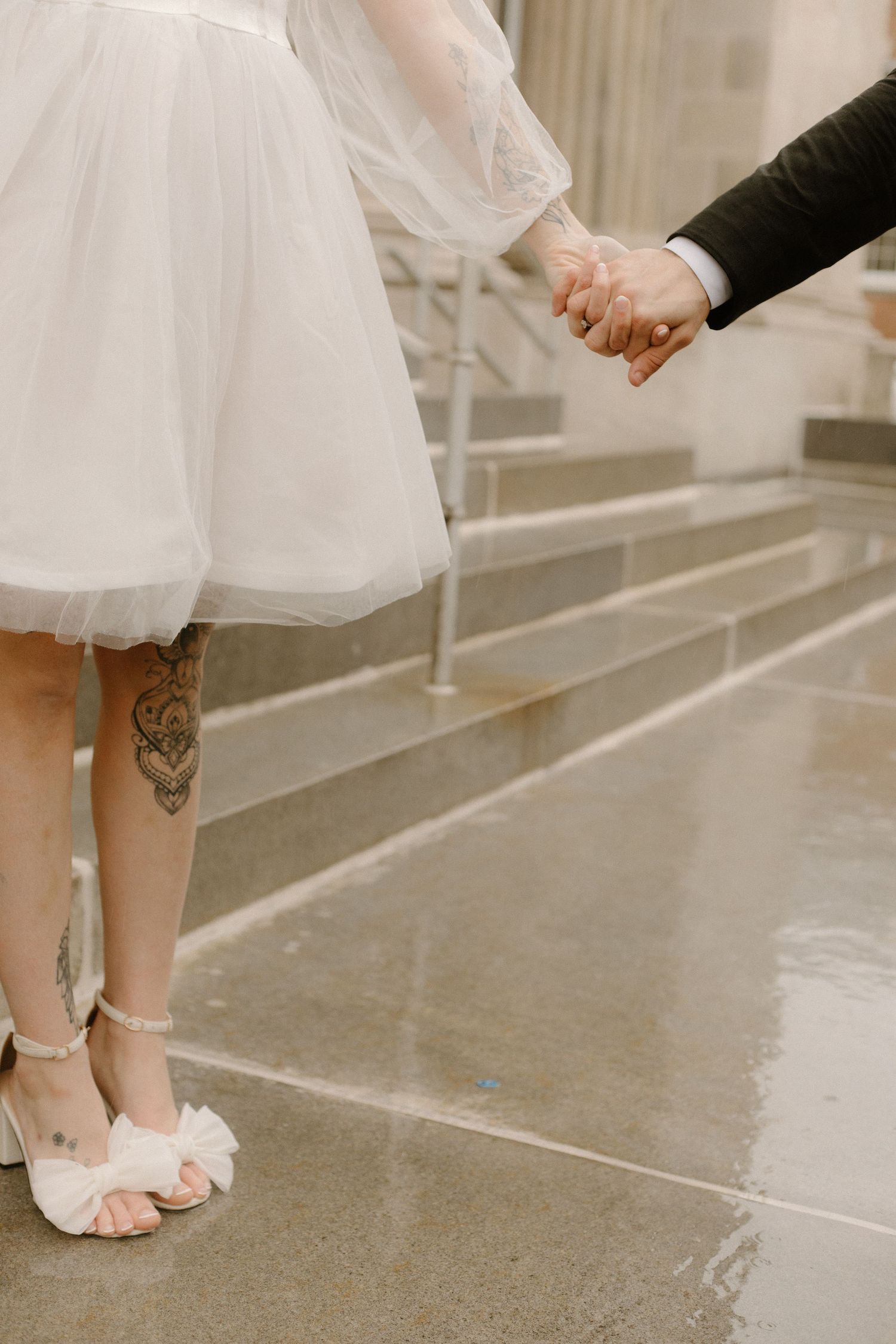 A tattooed leg in white heels and tulle dress holding hands with someone in a dark suit on stairs.