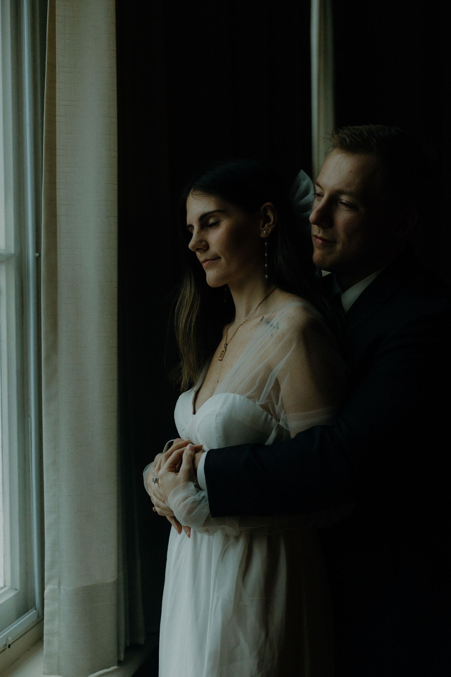 A romantic couple in formal attire standing by a window.