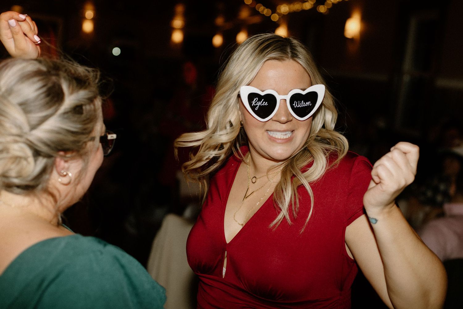 Person wearing heart-shaped sunglasses and red dress dancing at reception party.