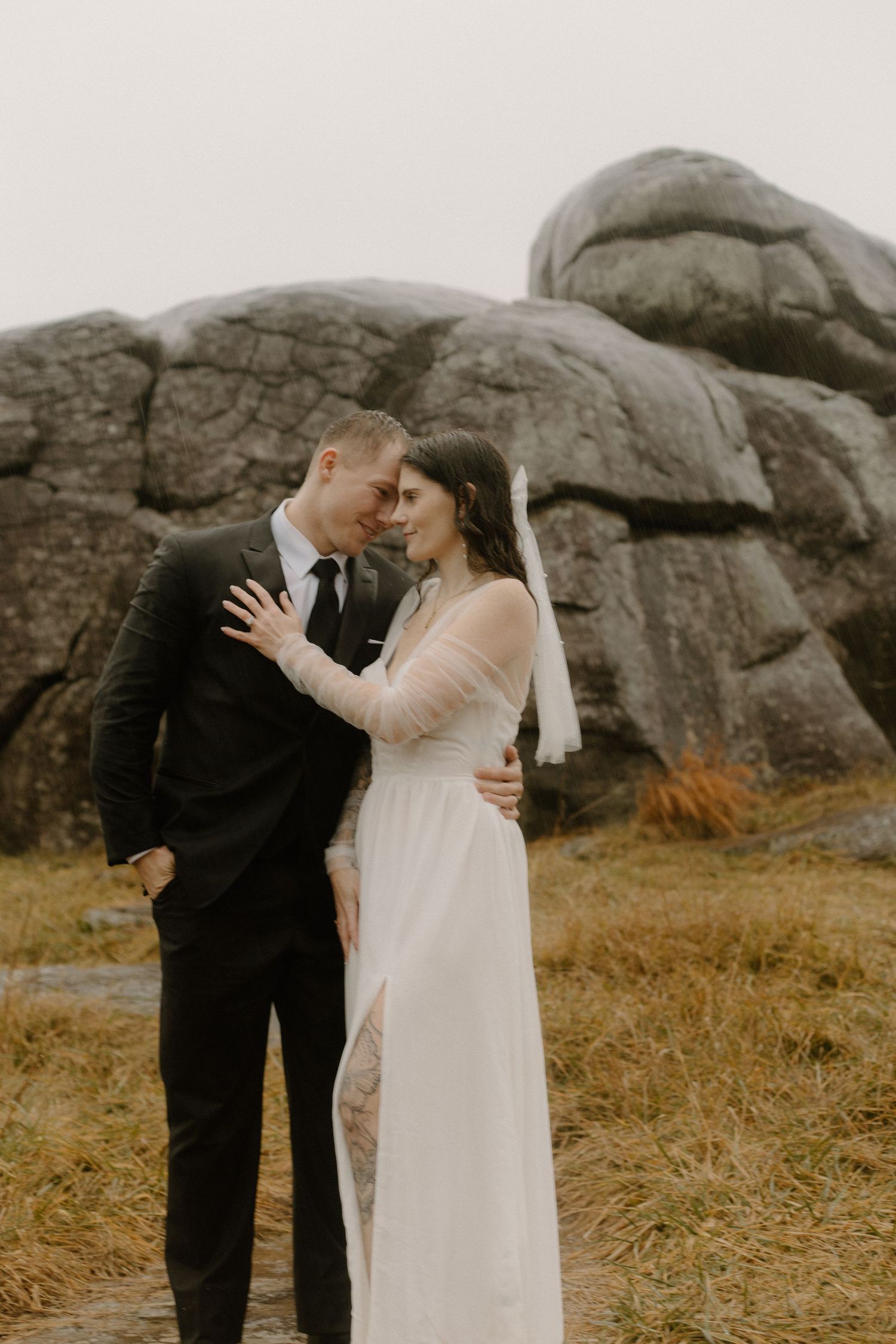 A couple in wedding attire sharing an intimate moment in front of large rock formations in a rustic outdoor setting.
